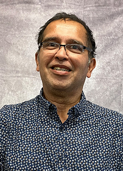 Man with short black hair wearing a navy and white button up shirt and narrow prescription glasses